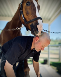 Man bending under a chestnut horse’s head, both sharing a gentle moment in a stable